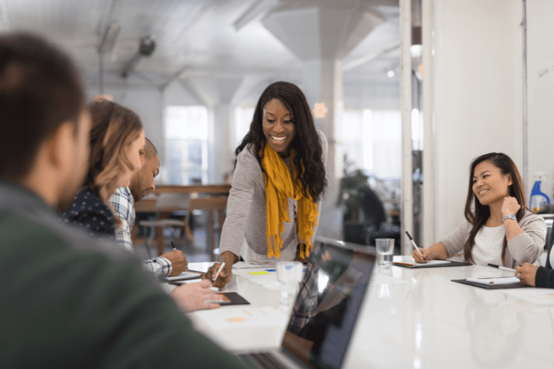 black woman with long brown hair and yellow scarf leading table discussion in office